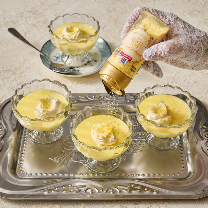 A hand in a lace glove pours a golden sauce from a bottle onto individual servings of custard topped with whipped cream and citrus zest, likely a dessert offering from the brand featured on the bottle.