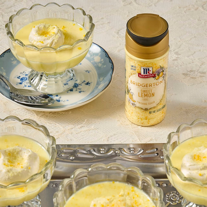 Three glass bowls filled with lemon pudding topped with whipped cream and lemon zest are displayed next to a bottle of McCormick Lemon &amp; Herb seasoning.