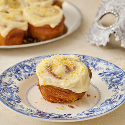 A frosted cinnamon roll sits on a blue and white patterned plate, with a platter of similar rolls and a decorative mask in the background, suggesting a festive occasion.