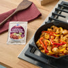 A packet of McCormick Chicken Fajita Skillet Sauce sits next to a cast iron skillet filled with cooked chicken and bell peppers on a stovetop.