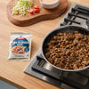A packet of Maggi Fix fA?r Chili con Carne sits next to a pan of cooked ground beef, with shredded lettuce, tomatoes, and a bowl of sauce in the background, suggesting a meal preparation scene.