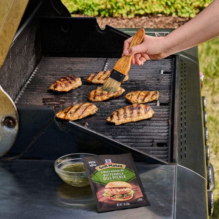 A person uses a basting brush to apply a herb-infused marinade to chicken breasts grilling on a barbecue next to a packet of Mccormick Grill Mates Buttermilk Dill Pickle Seasoning Mix, 1.0 Oz.