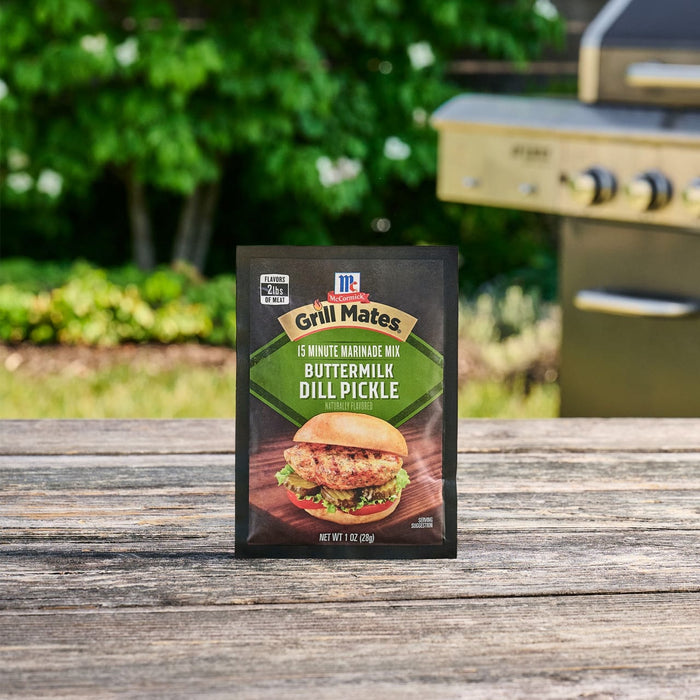 A packet of Mccormick Grill Mates Buttermilk Dill Pickle Seasoning Mix, 1.0 Oz stands on a rustic wooden table with a grill and green foliage in the background.