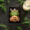 A packet of Mccormick Grill Mates Buttermilk Dill Pickle Seasoning Mix, 1.0 Oz is centered on a dark textured surface surrounded by fresh dill sprigs and a jar of buttermilk.