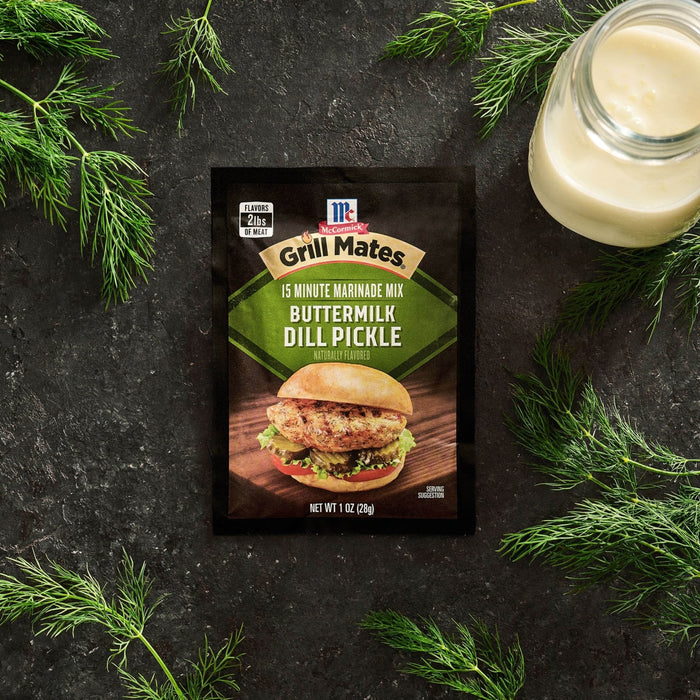 A packet of Mccormick Grill Mates Buttermilk Dill Pickle Seasoning Mix, 1.0 Oz is centered on a dark textured surface surrounded by fresh dill sprigs and a jar of buttermilk.