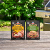 Two packets of McCormick Grill Mates 15 Minute Marinade Mix in Tennessee Style Whiskey Honey and Mccormick Grill Mates Buttermilk Dill Pickle Seasoning Mix, 1.0 Oz sit on a rustic wooden table in front of a grill and lush green foliage.