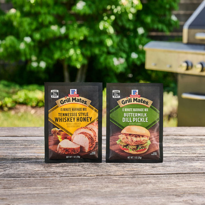 Two packets of McCormick Grill Mates 15 Minute Marinade Mix in Tennessee Style Whiskey Honey and Mccormick Grill Mates Buttermilk Dill Pickle Seasoning Mix, 1.0 Oz sit on a rustic wooden table in front of a grill and lush green foliage.