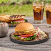 A delicious-looking grilled chicken sandwich is served on a rustic plate with a side of red potato salad, with another sandwich and two glasses of cola in the background for an outdoor meal.