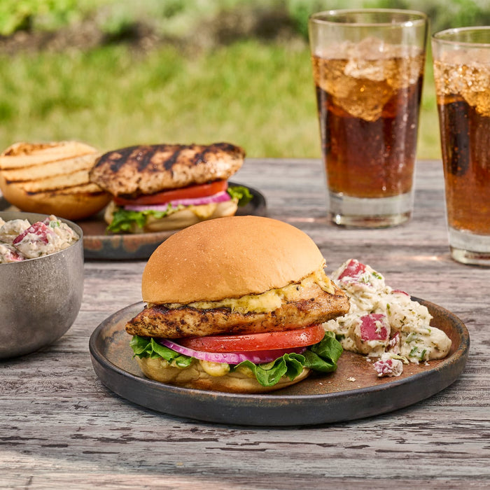A delicious-looking grilled chicken sandwich is served on a rustic plate with a side of red potato salad, with another sandwich and two glasses of cola in the background for an outdoor meal.