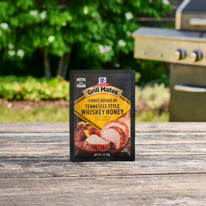 A package of Mccormick Grill Mates Tennessee Style Whiskey Honey Marinade Seasoning Mix, 1.0 Oz stands on a rustic wooden table with a grill and green foliage in the background.