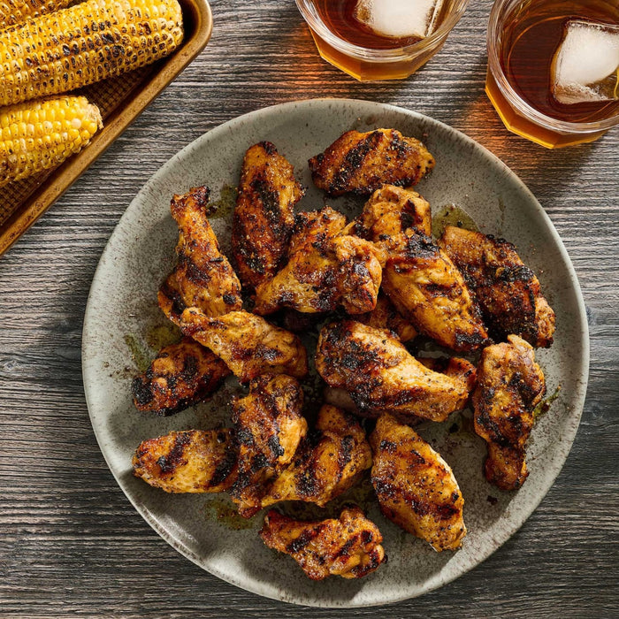 A plate of grilled chicken wings seasoned with Mccormick Grill Mates Tennessee Style Whiskey Honey Marinade Seasoning Mix 1.0 Oz served alongside charred corn on the cob and iced drinks.