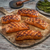 Five grilled salmon fillets with crosshatch grill marks are arranged on a rustic wooden cutting board, with a side of grilled asparagus and a small bowl of honey in the background.