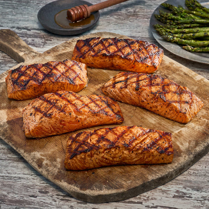 Five grilled salmon fillets with crosshatch grill marks are arranged on a rustic wooden cutting board, with a side of grilled asparagus and a small bowl of honey in the background.