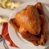 A whole roasted turkey seasoned with Mccormick Turkey Dry Brine Rub, 12.2 Oz sits on a white platter next to a glass of white wine.