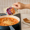 A hand pours the contents of a Mccormick Spicy Vodka Sauce Seasoning Mix, 1.0 Oz packet into a blue pan containing a creamy, reddish-orange sauce.