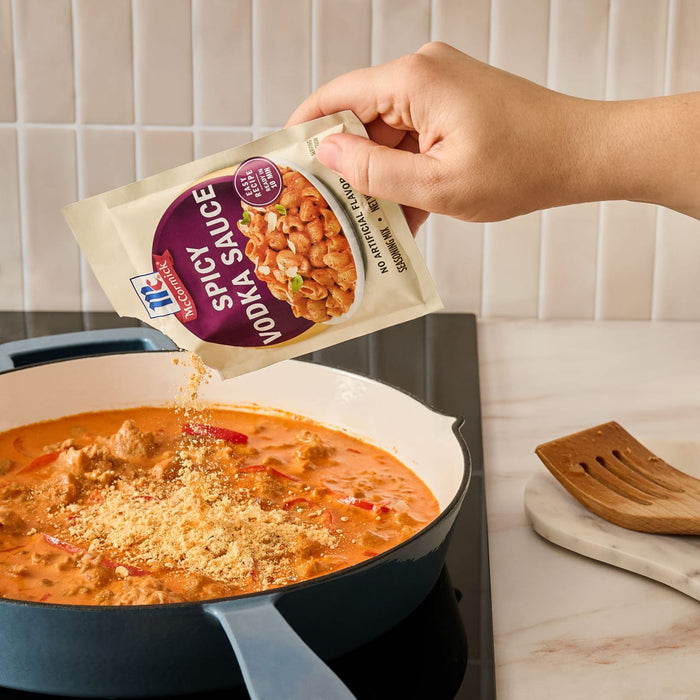 A hand pours the contents of a Mccormick Spicy Vodka Sauce Seasoning Mix, 1.0 Oz packet into a blue pan containing a creamy, reddish-orange sauce.