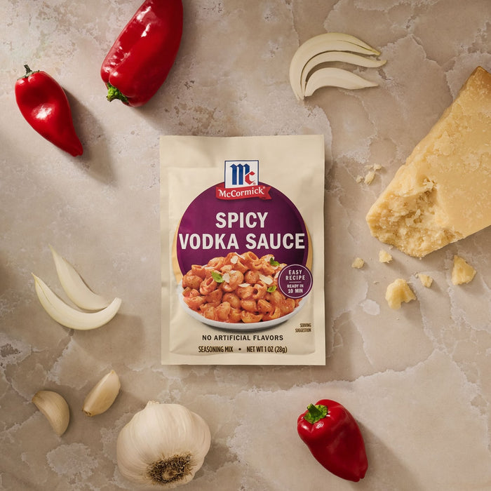 A packet of Mccormick Spicy Vodka Sauce Seasoning Mix, 1.0 Oz, is displayed on a marble countertop surrounded by fresh ingredients like red peppers, garlic, onion, and a wedge of Parmesan cheese.
