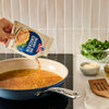 A hand pours the contents of a packet of Mccormick Marry Me Chicken Seasoning Mix, 0.75 Oz into a blue pan filled with broth on a stovetop.