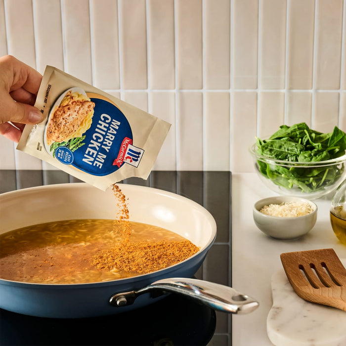A hand pours the contents of a packet of Mccormick Marry Me Chicken Seasoning Mix, 0.75 Oz into a blue pan filled with broth on a stovetop.