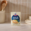 A packet of Mccormick Marry Me Chicken Seasoning Mix, 0.75 Oz, is displayed upright on a light-colored kitchen countertop, with a utensil holder and a stack of plates visible in the background.