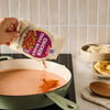 A hand pours the contents of a Mccormick Chicken Tikka Masala Seasoning Mix, 0.87 Oz packet into a light green pan of creamy sauce, demonstrating an easy way to prepare the dish.