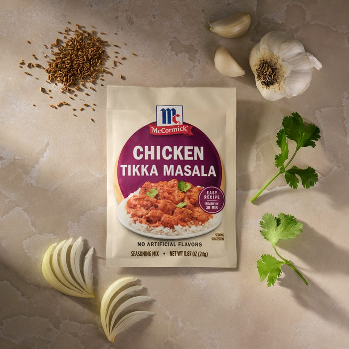 A packet of Mccormick Chicken Tikka Masala Seasoning Mix, 0.87 Oz is arranged on a countertop with fresh ingredients like garlic, onion, cilantro, and cumin seeds.