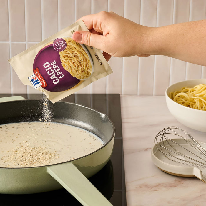 A hand pours the contents of a packet of Mccormick Cacio E Pepe Seasoning Mix, 1.12 Oz into a light green pan with a creamy sauce to prepare a meal.
