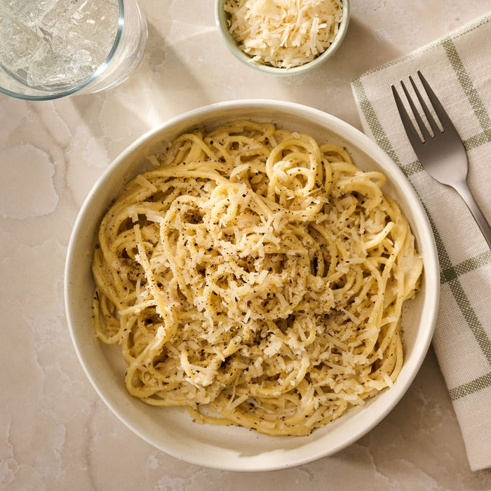 An overhead shot shows a bowl of spaghetti prepared with Mccormick Cacio E Pepe Seasoning Mix, 1.12 Oz, topped with grated cheese and black pepper, next to a glass of water and a fork.