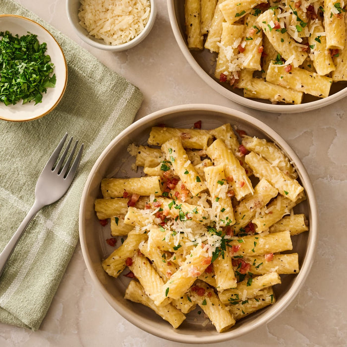 Two bowls of rigatoni pasta, prepared using Mccormick Cacio E Pepe Seasoning Mix, 1.12 Oz, are garnished with cheese, parsley, and bacon bits on a marble countertop.