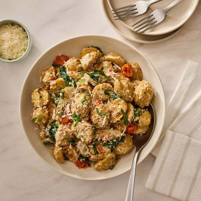 A large bowl of creamy gnocchi with spinach and tomatoes, prepared with Mccormick Cacio E Pepe Seasoning Mix, 1.12 Oz, is served on a marble countertop next to plates and a bowl of parmesan cheese.