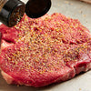 A raw steak is being seasoned with spices from a shaker.