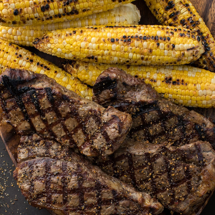 Grilled steaks with distinct grill marks are paired with grilled corn on the cob, suggesting a summer barbecue.