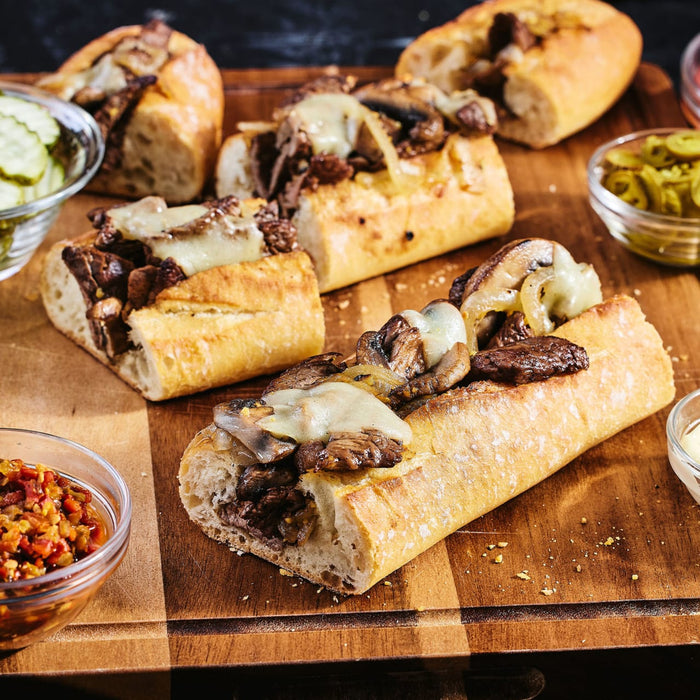 A wooden board displays several cheesesteak sandwiches topped with mushrooms and cheese, accompanied by bowls of pickles and other condiments.