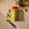 An overhead shot features a bottle of Mccormick Garlic And Olive Oil Seasoning Blend, 5.25 Oz, surrounded by its core ingredients: fresh garlic cloves, sprigs of rosemary and thyme, and a cruet of olive oil.