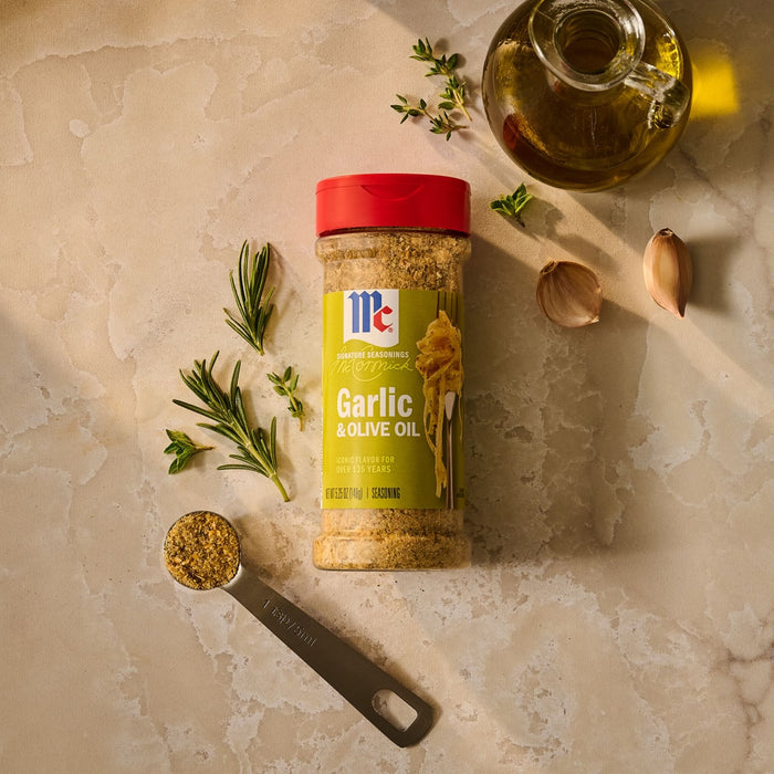 An overhead shot features a bottle of Mccormick Garlic And Olive Oil Seasoning Blend, 5.25 Oz, surrounded by its core ingredients: fresh garlic cloves, sprigs of rosemary and thyme, and a cruet of olive oil.