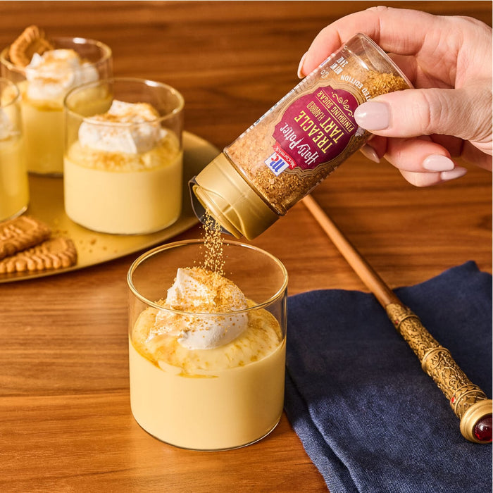 A hand sprinkles Mccormick Harry Potter Treacle Tart Finishing Sugar, 3.43 Oz onto a pudding dessert topped with whipped cream.