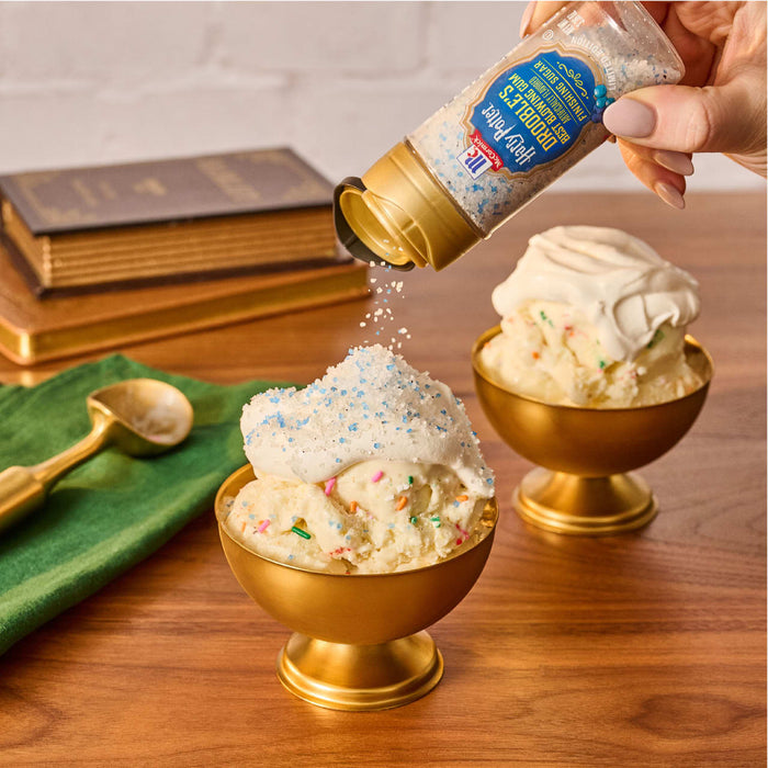 A hand sprinkles Mccormick Harry Potter Drooble'S Best Blowing Gum Finishing Sugar, 3.36 Oz onto a golden bowl of vanilla ice cream topped with whipped cream.