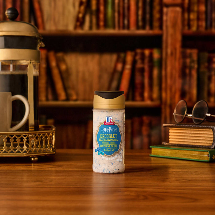 A bottle of Mccormick Harry Potter Drooble'S Best Blowing Gum Finishing Sugar, 3.36 Oz, sits on a wooden desk in a cozy library setting, complete with a French press, a mug, and round glasses resting on old books.