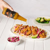A hand pours hot sauce from a bottle onto shrimp tacos, with a side of slaw and avocado slices.