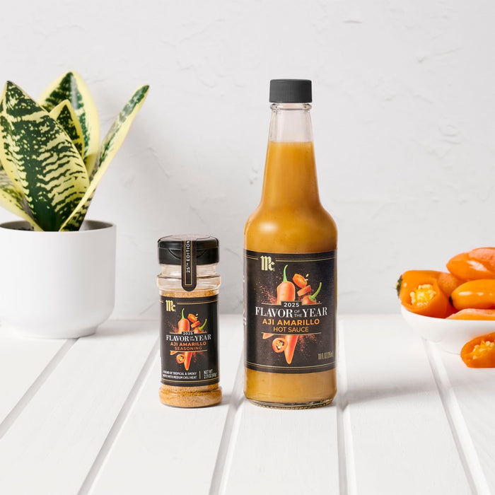 A bottle of Yellow Fruit hot sauce and a jar of Yellow Fruit seasoning are displayed on a white table with a plant and peppers.