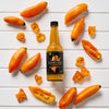 A bottle of Clark and Hopkins Artisan Pepper Sauce is surrounded by fresh, vibrant yellow peppers on a white wooden surface.