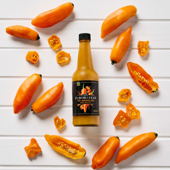 A bottle of Clark and Hopkins Artisan Pepper Sauce is surrounded by fresh, vibrant yellow peppers on a white wooden surface.