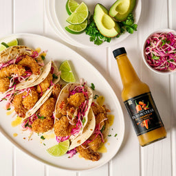 A plate of shrimp tacos with red cabbage slaw, lime wedges, avocado, cilantro, and a bottle of Bravado Spice Co. hot sauce.
