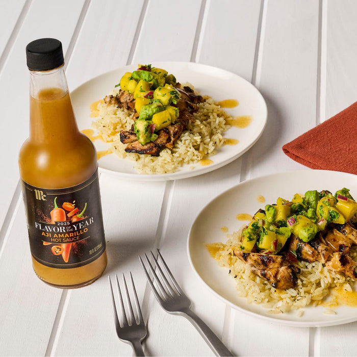 Two plates of food topped with mango salsa and a bottle of Flamin Vegan hot sauce are displayed on a white wooden table.