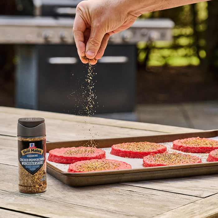 A hand sprinkles Mccormick Grill Mates Peppercorn And Worcestershire Seasoning Cracked 2.75 Oz onto raw hamburger patties on a baking sheet, with a grill in the background.