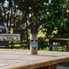 A bottle of Mccormick Grill Mates Peppercorn And Worcestershire Seasoning Cracked 2.75 Oz sits on a wooden patio table with a grill and lush greenery in the background.