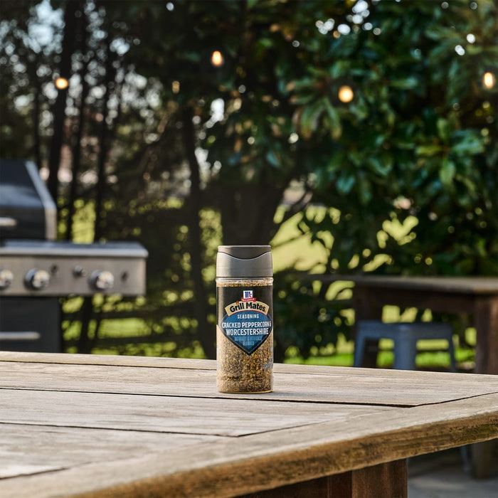 A bottle of Mccormick Grill Mates Peppercorn And Worcestershire Seasoning Cracked 2.75 Oz sits on a wooden patio table with a grill and lush greenery in the background.