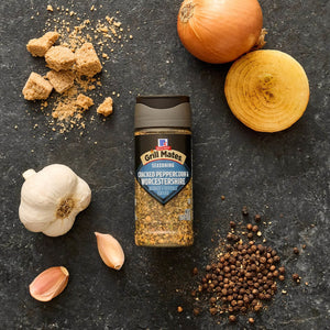 A bottle of McCormick Grill Mates Cracked Peppercorn &amp; Worcestershire seasoning is surrounded by garlic, brown sugar, black peppercorns, and onion on a dark surface.