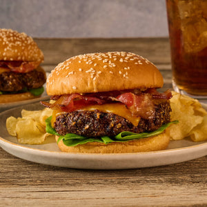 A bacon cheeseburger with lettuce and potato chips is served on a plate, with a second burger and a glass of iced tea in the background.