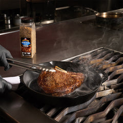 A cast iron skillet on a gas stove griddle with a steak cooking, and a bottle of McCormick's Steak Seasoning in the background. A person wearing black gloves is using tongs to handle the skillet.